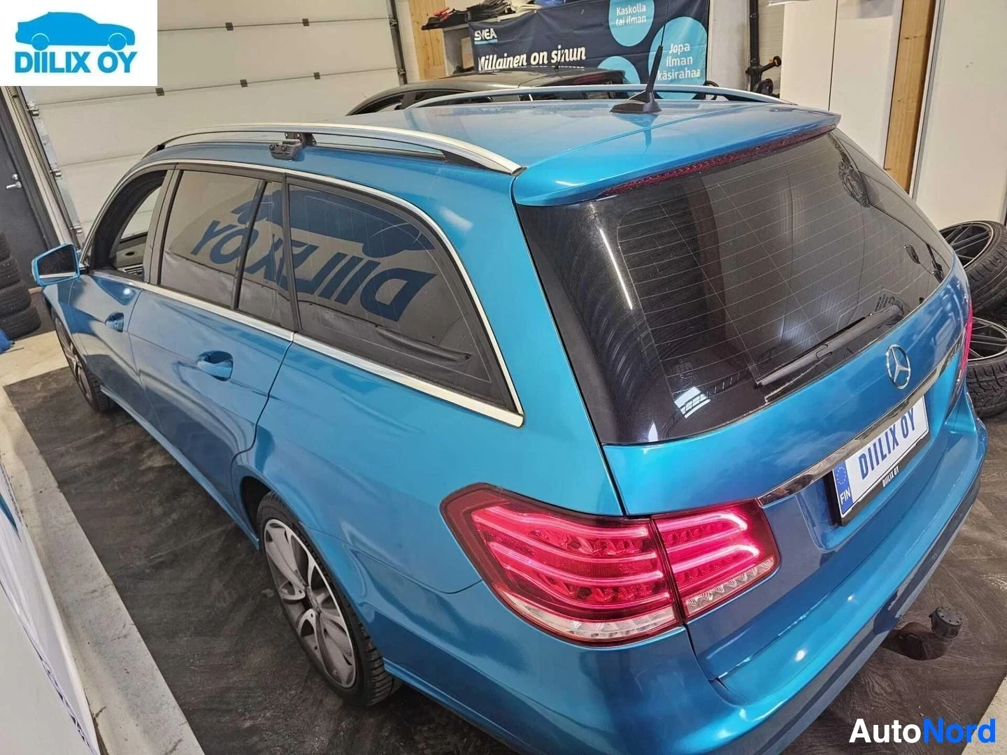 Takaviisto: Sininen Mercedes-Benz estate-auto näkyy takaa ja sivulta sisäkattohyllyllä ja punaisilla takavaloilla. Ajoneuvo on sisätiloissa betonilattian päällä.