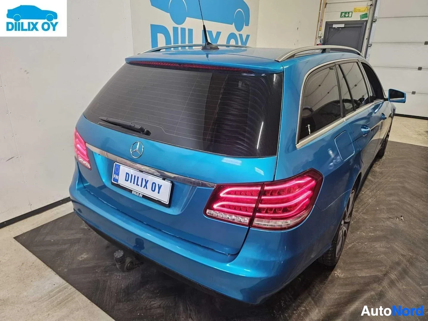 Takaa: Sininen Mercedes-Benz farkkuauto sisätiloissa, takaosasta kuvattuna. Näkyvät takavalot, rekisterikilpi ja tavaratilan ikkuna.