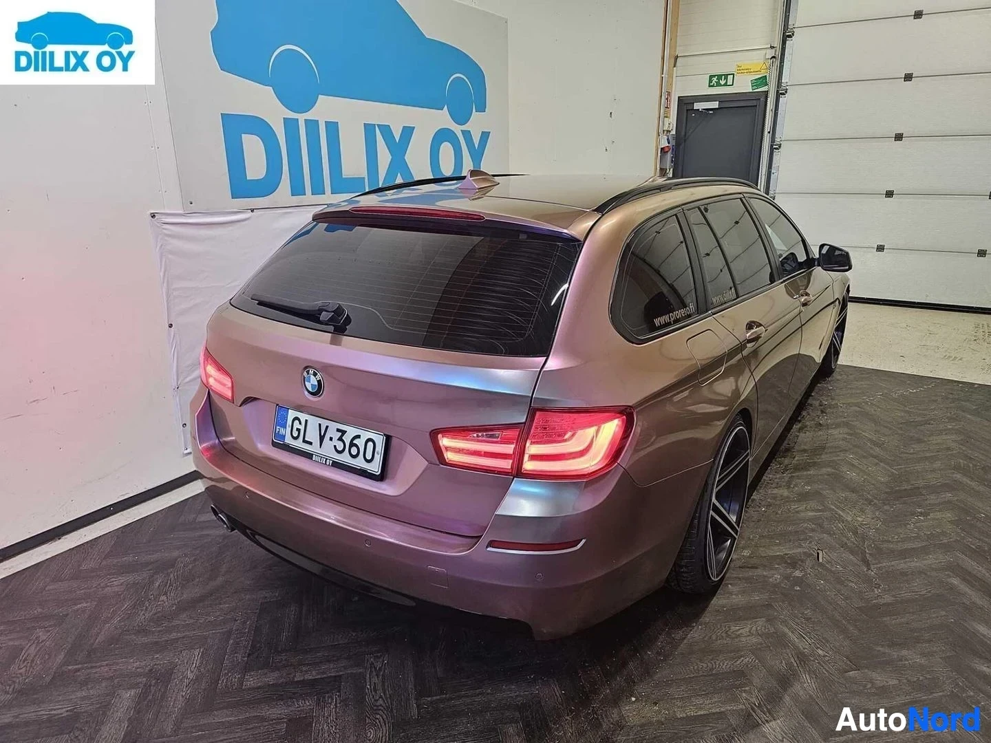 Takaviisto: Violetti BMW farmari, takaosan oikea kulma, sisätiloissa. Rekisterikilpi näkyy. Punaiset takavallot palavat. Diilix Oy:n seinämainonta taustalla