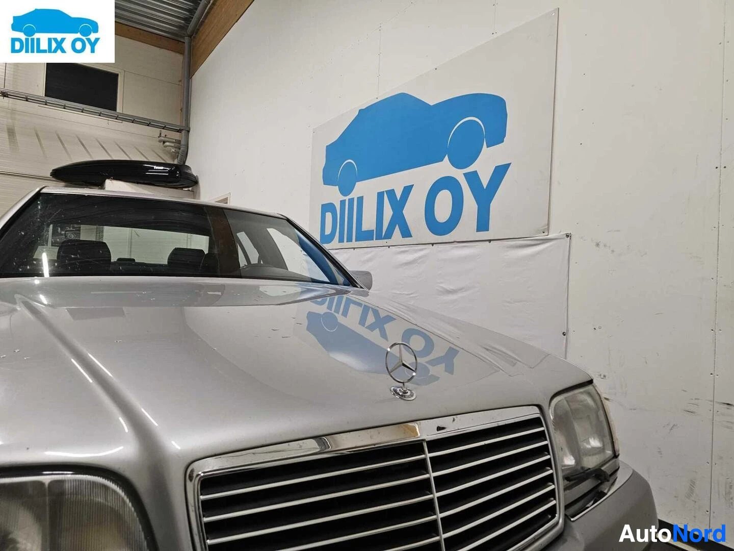 Edestä: Valkoisen Mercedes-Benzin etupuoli yksityiskohta. Säleikkögrilli, etuvalaisimet ja Mercedes-Benz-logo kojelaudalla näkyvät. Varaston seinä taustalla.