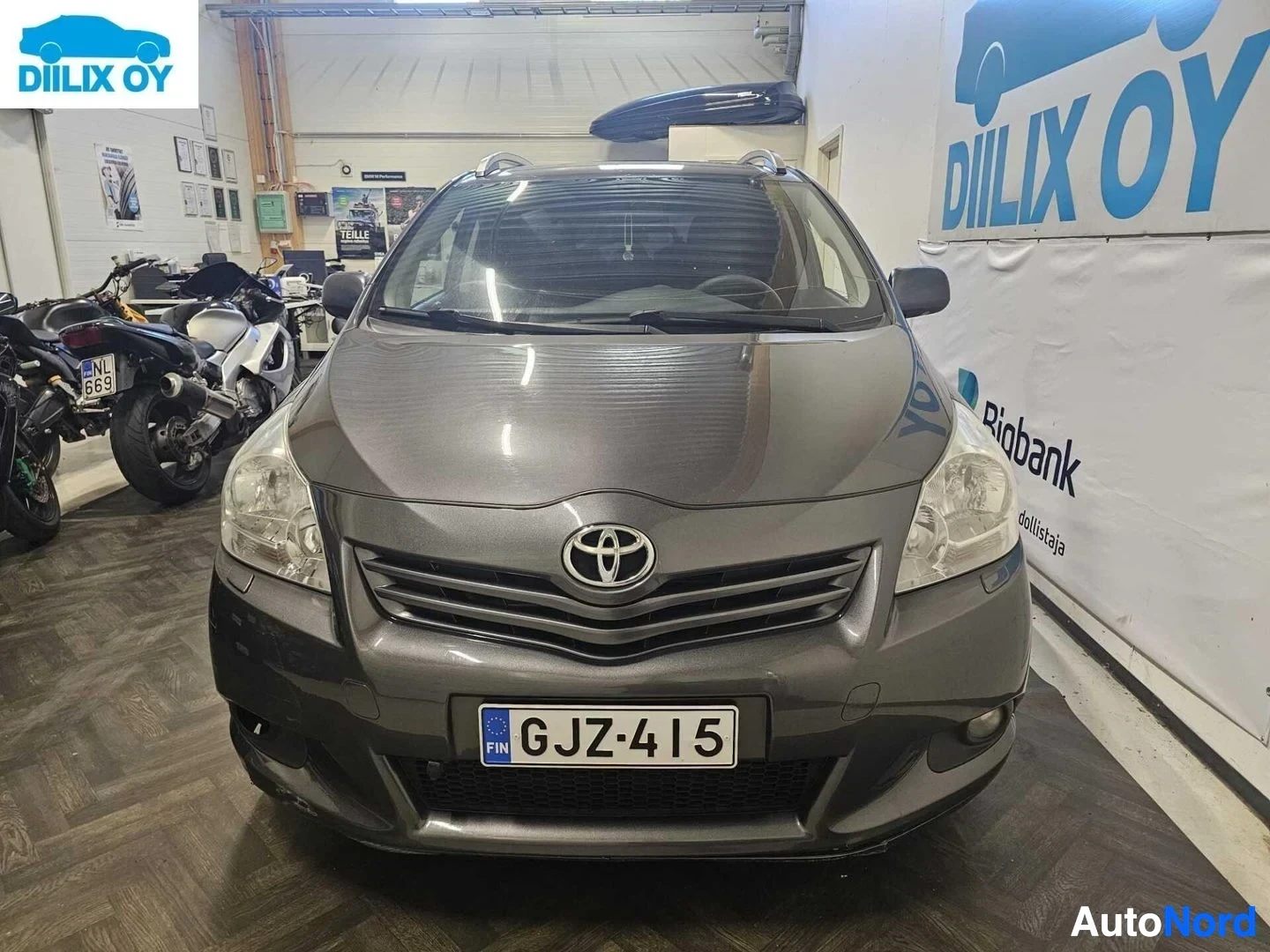 Edestä: Harmaa Toyota Verso suoraan edestä kuvattuna hallissa, rekisterinumero näkyvissä