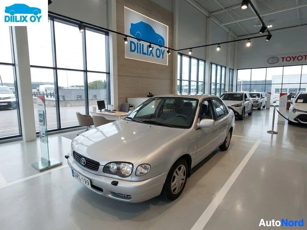 Etuviisto, nokka vasemmalle: Valkoinen Toyota Corolla neliovinen sedan sisätiloissa autoliikkeellä, etupuoli näkyy vasemmalle kallistuneena, rekisterikilvessä näkyy teksti.