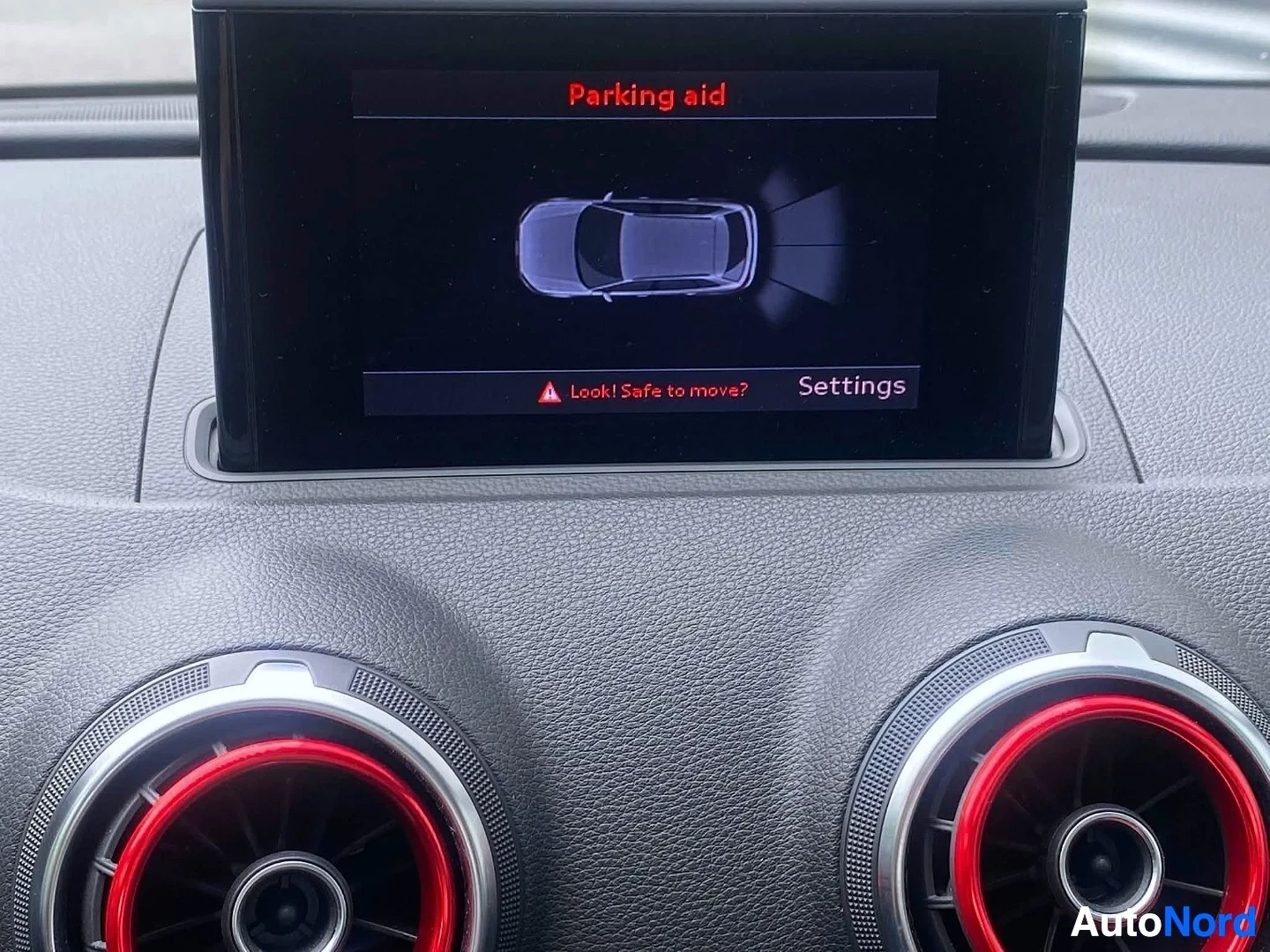 Ohjaamo: Näyttö näyttää paikantutkan näkymää ylhäältä katsottuna. Punaisia ympyrä-muotoisia painikkeita näkyvät alla ja näytön yläosassa teksti 'Parking aid'.