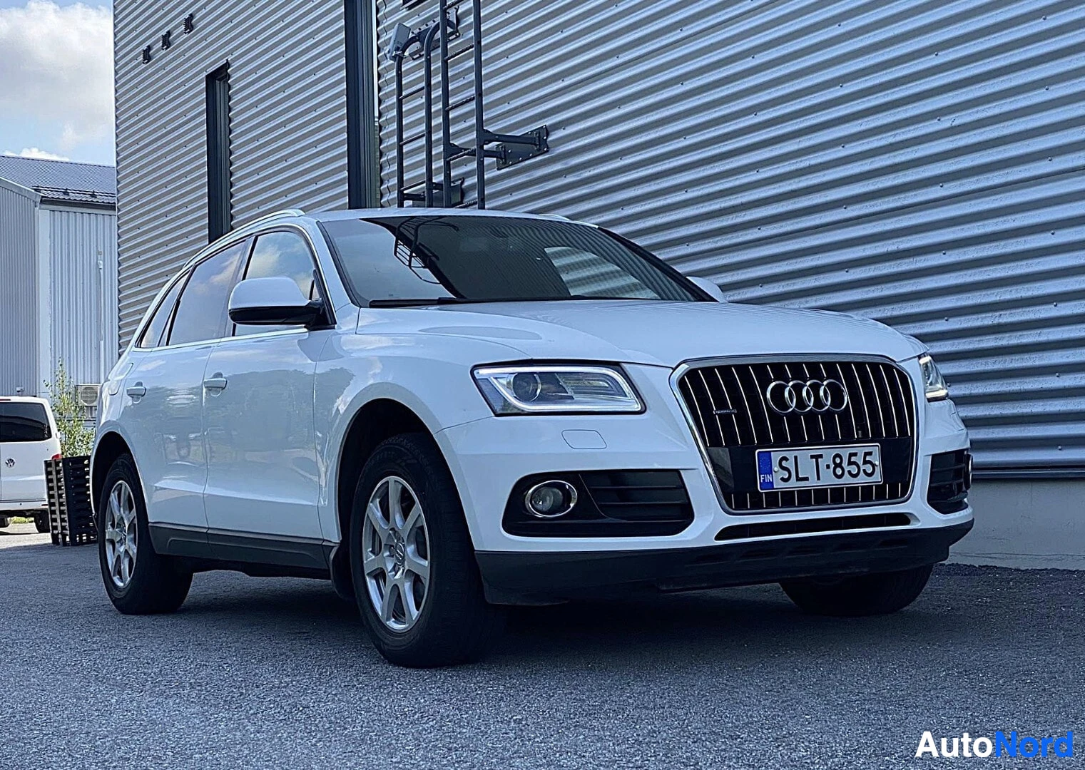 Etuviisto, nokka oikealle: Valkoinen Audi Q5 -maastoauto vasemmasta etukolmesta, rekisterinumero näkyvissä, teräslevyseinä taustalla