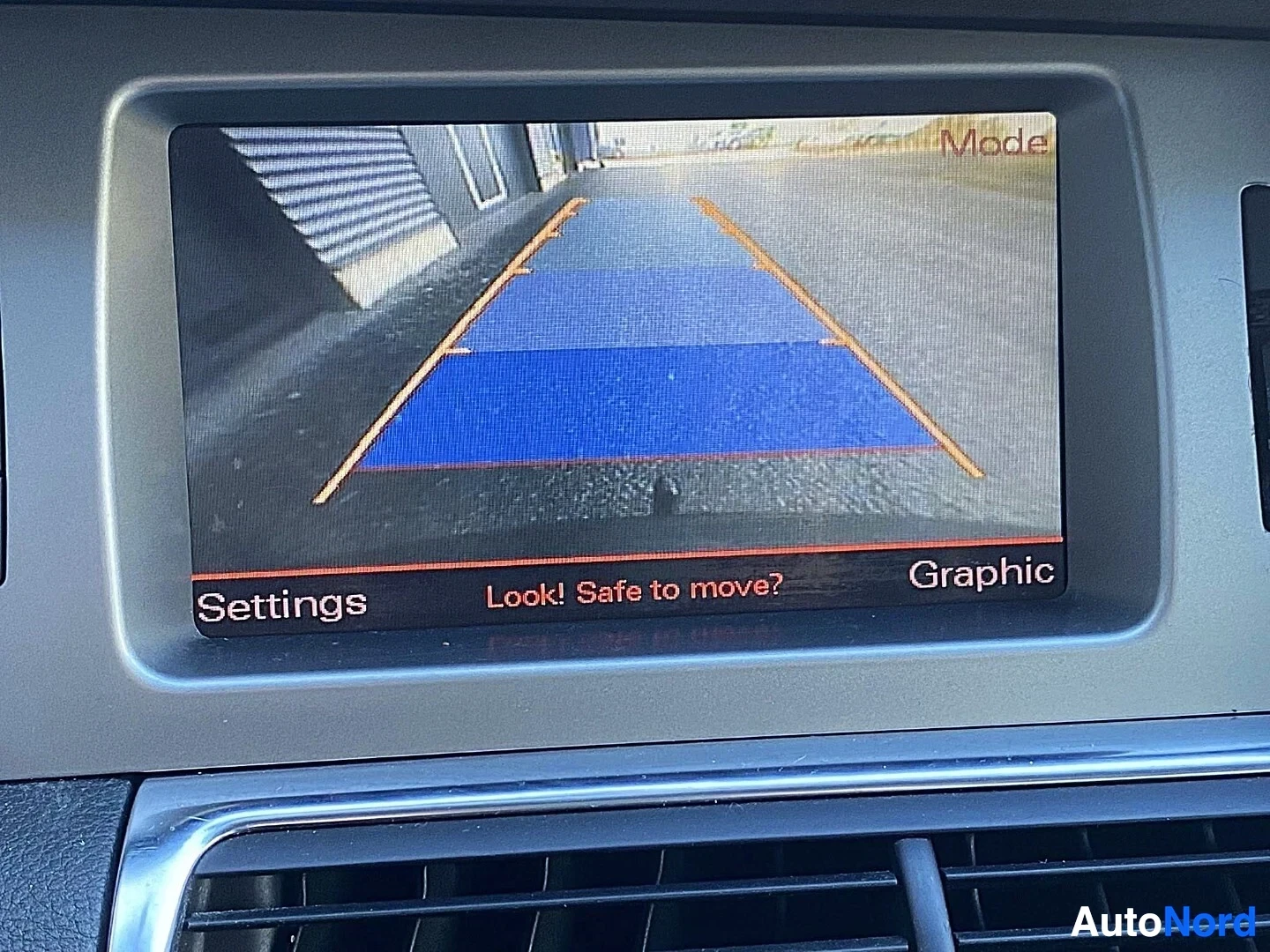Yksityiskohta: Navigointiruudukon näyttö kameran takakuvassa, harmaa ajoradan kuva ja sininen turvavyöhyke, tekstit Mode, Settings, Look! Safe to move?, Graphic näkyvissä.