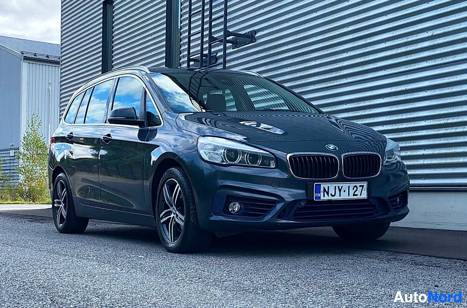 Etuviisto, nokka vasemmalle: Sininen BMW-tila-auto näkyy etulta hieman vasemmalta, pysäköitynä korrugoidun metallirakennuksen eteen. Rekisterikilpi NJY·127 näkyvissä.