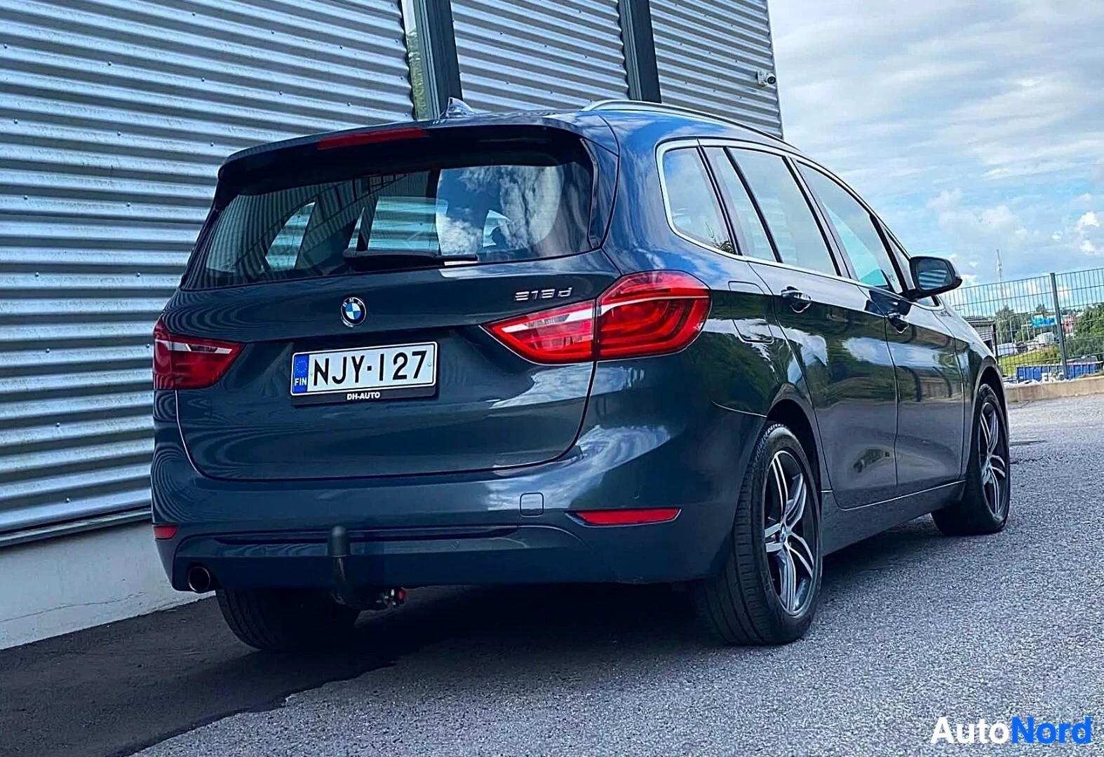 Takaviisto: Sinisen BMW-tila-auton takapuoli näkyvissä oikealta, punaisia takavaloja ja rekisterikilpeä NJY·127 näyttäen. Merkki '218d' näkyy takalisäkkeessä.