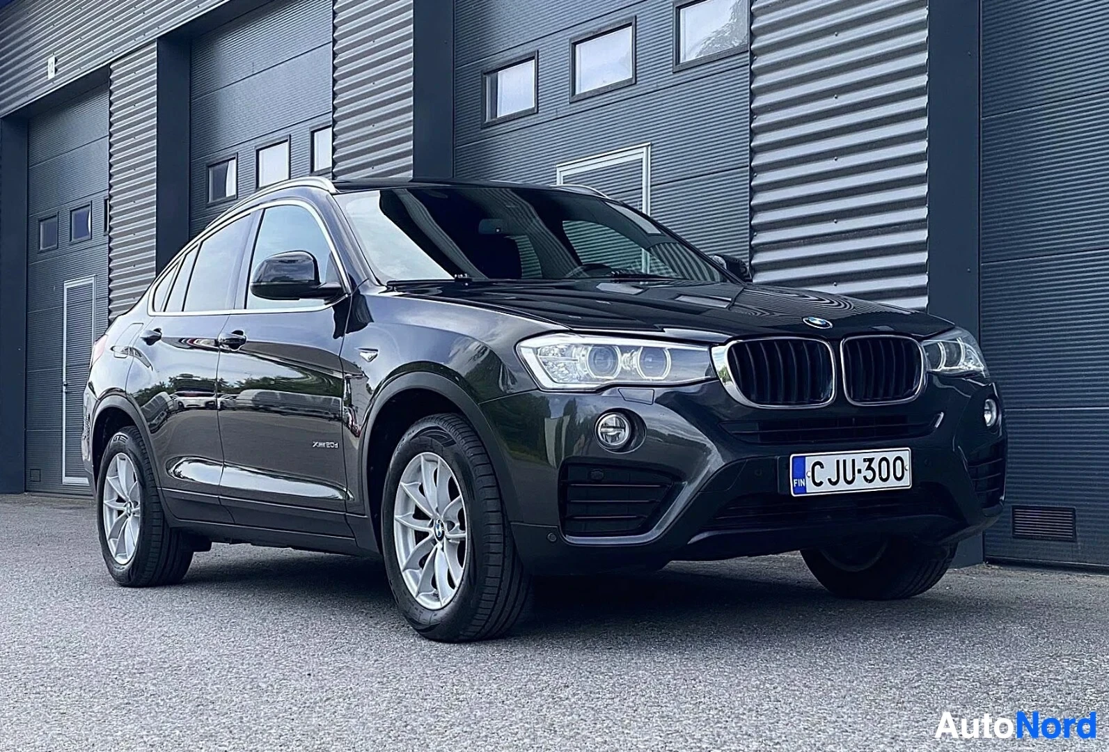 Etuviisto, nokka oikealle: Musta BMW X4 edestä ja sivusta katsottuna, rekisterinumero näkyvissä, valkeat metalliset vanteet, teollisuusrakennuksen seinä taustalla