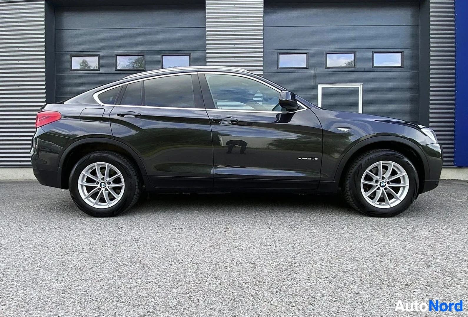 Sivulta: Musta BMW X4 F26 näkyy sivusta vasemmalla olevalla polkimenä oikean suuntaan, valkoisten aluepyörien ja punaisen takaliidin kanssa harmaalla asfaltin edessä teo