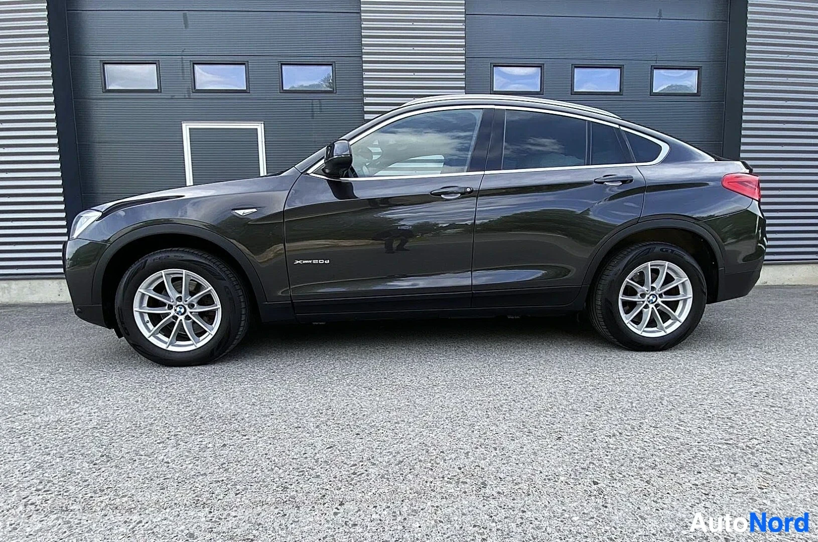 Sivulta: Musta BMW X4 F26 sivusta vasemmalla, valkoisten aluepyörien kanssa, ovi auki, harmaalla asfaltin edessä teollisuusrakennusta vasten.