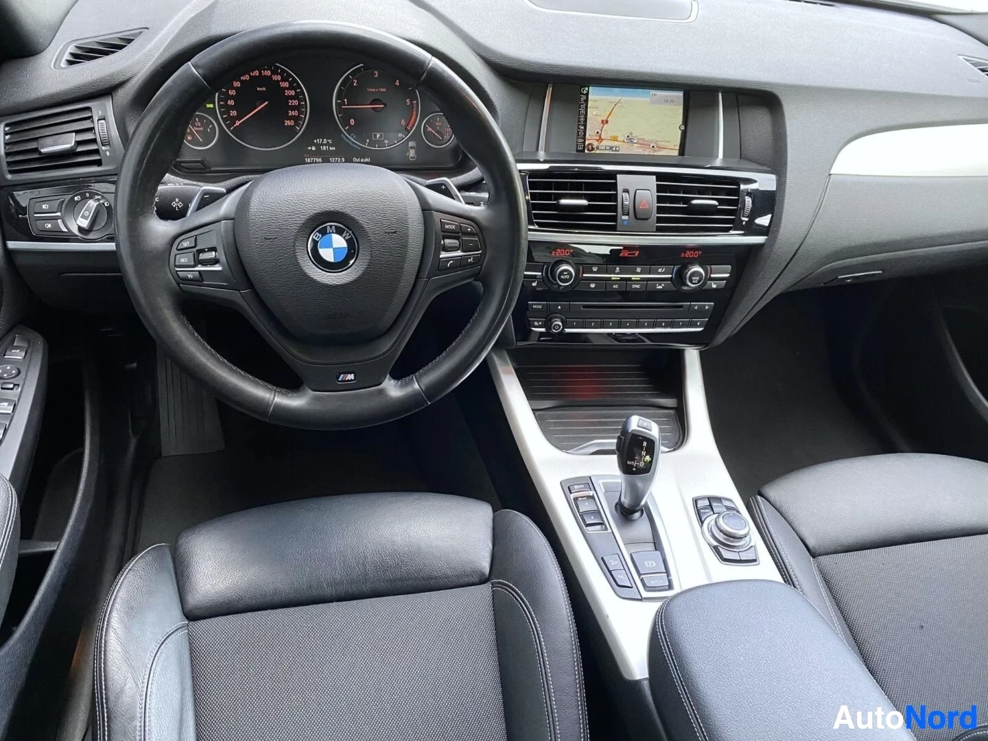Ohjaamo: BMW-ohjauksinen mustan nahkaisen ohjauspyörän yläpuolella kojelauta näyttöineen, keskellä navigointilaite kartalla, auton etuosassa käyttöpaneeli ja istuminen.