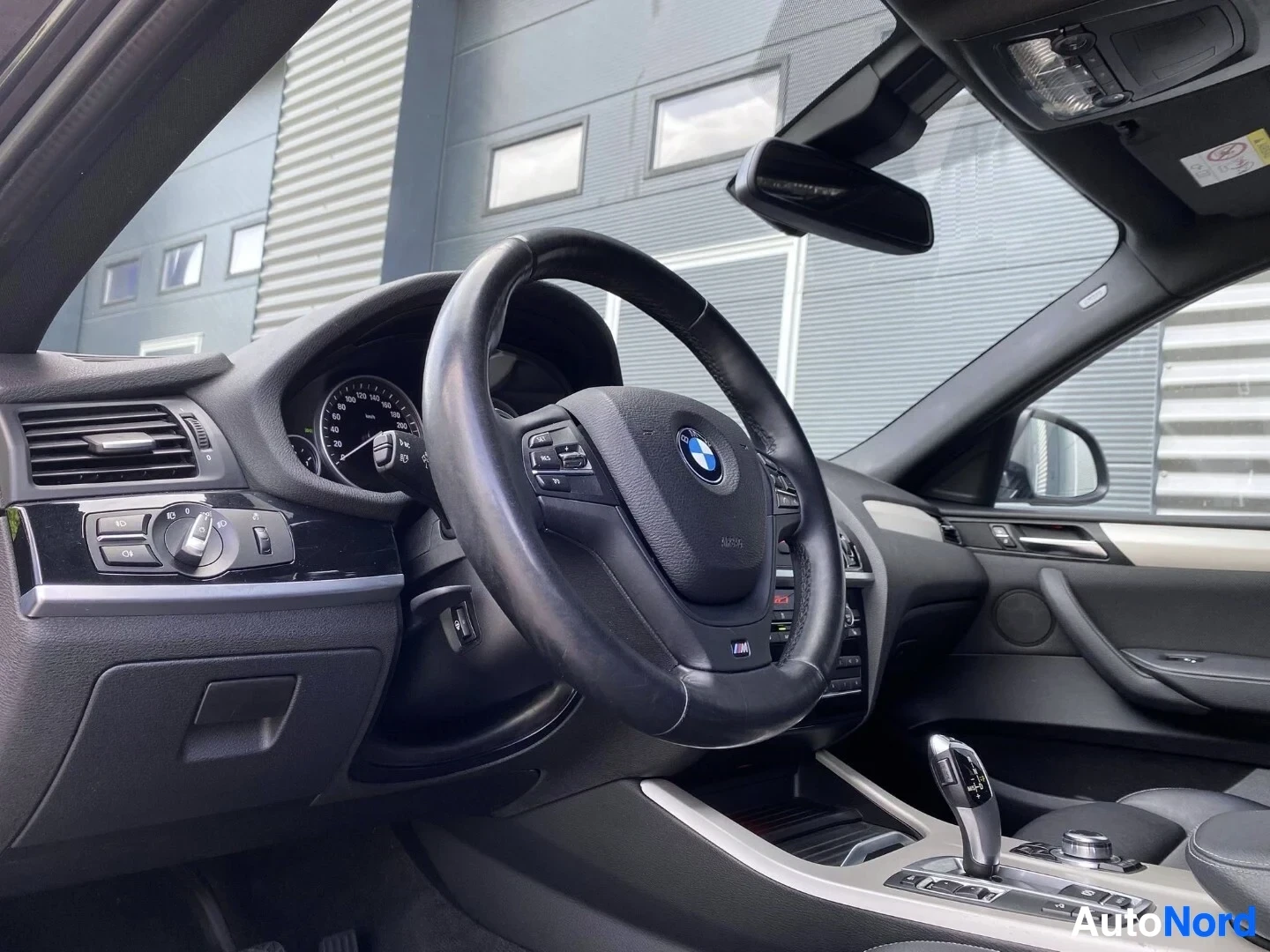 Yksityiskohta: Lähikuva ohjauspyörästä ja instrumenttipaneelista; näkyy BMW-merkki, mustainen nahkainen ohjauspyörä, digitaaliset instrumentit, ja harmaita sisäänottomateriaal