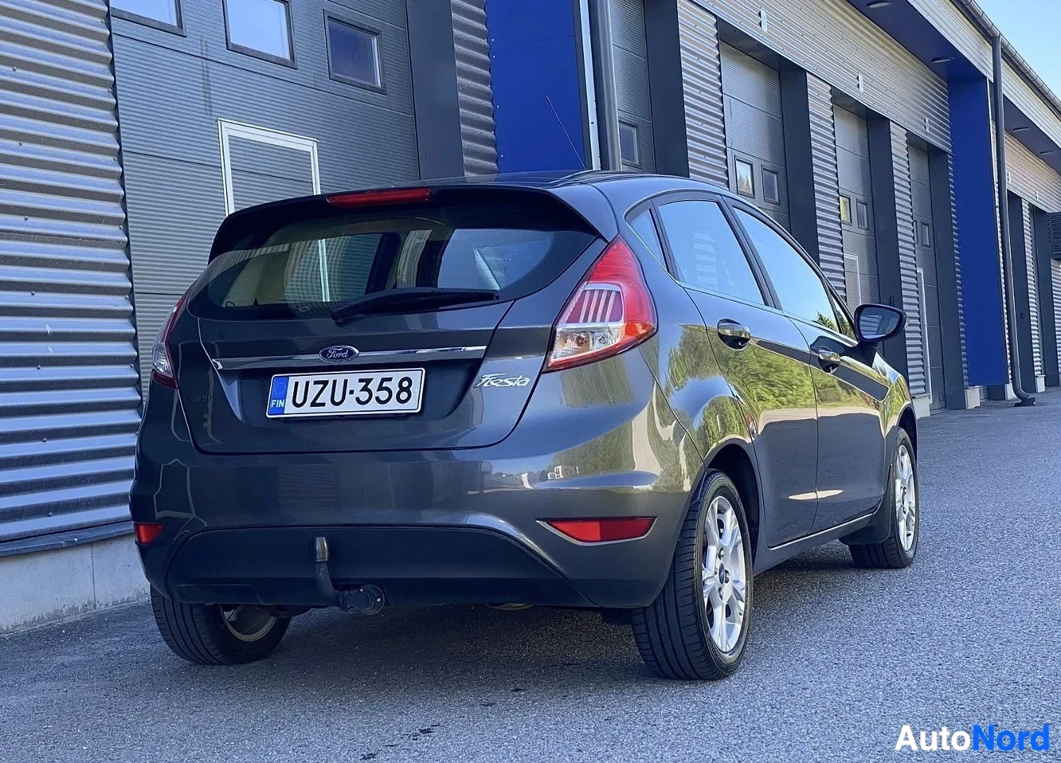 Takaa: Tummansininen Ford Fiesta takaa, punaisten takavalojen ja rekisterikylven näkyessä, teollisuushallien edessä
