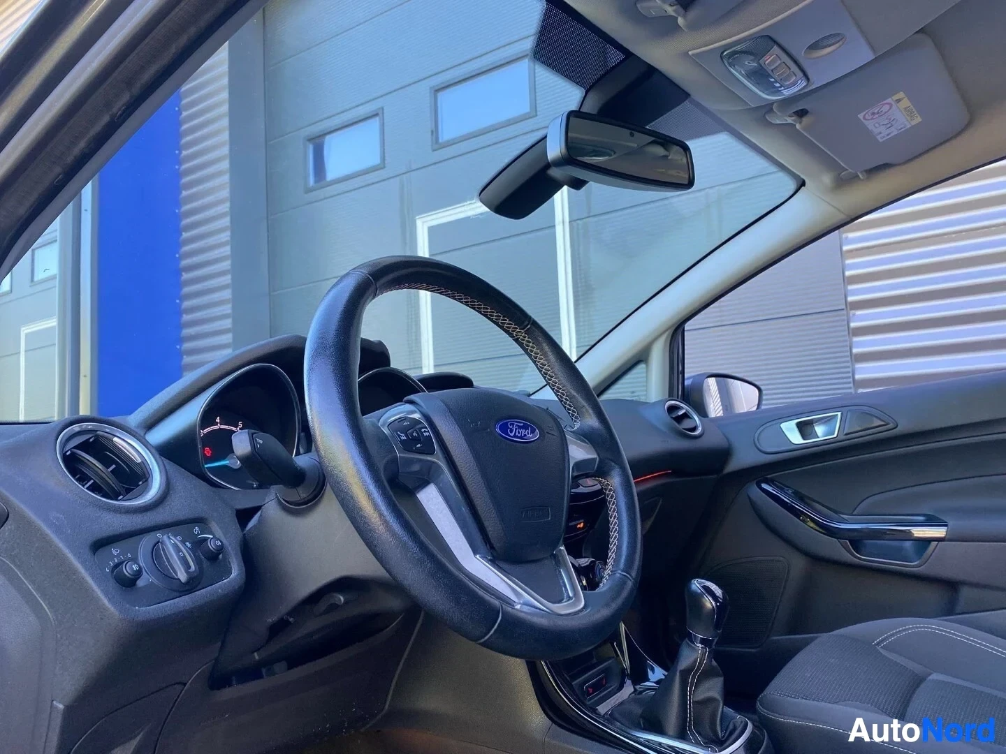 Ohjaamo: Ford-merkkinen ohjauspyörä ja kojelauta auton sisustuksesta, näkymä pois päin. Peiliä näkyy tuulilasissa, sisäkaton säilytyslokerot näkyvät yläpuolella.
