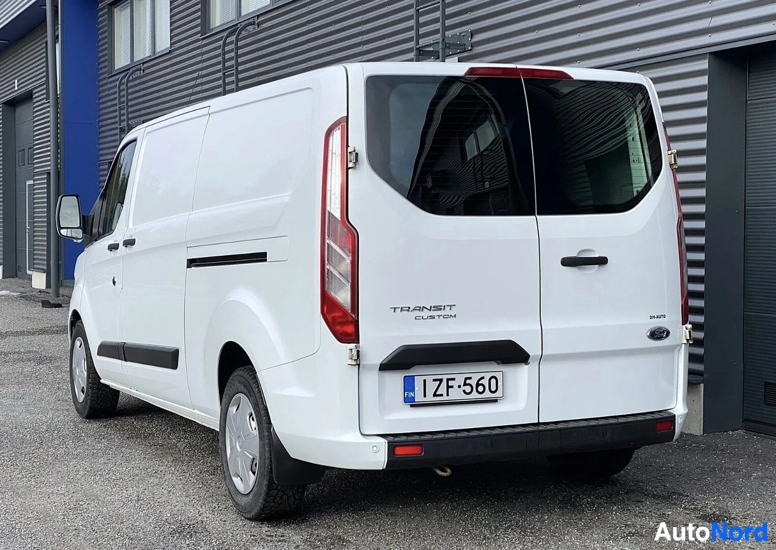 Takaviisto: Valkoinen Ford Transit Custom -pakettiauto kuvattu takaa vasemmalta puolelta, rekisterikilvessä, takaosassa näkyy TRANSIT CUSTOM ja DH AUTO -teksti