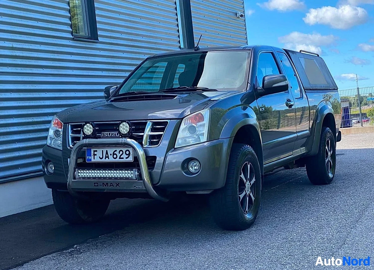 Etuviisto, nokka vasemmalle: Sininen Isuzu D-Max kaksiohjaamo kuorma-auto etuviistosta vasemmalle orientoituneella nenällä, metallisuojaputki edessä, rekisteritunnus