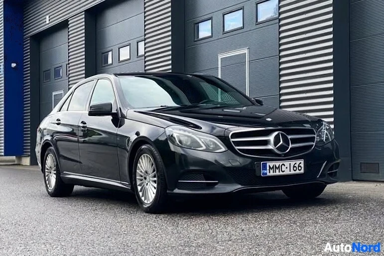 Etuviisto, nokka vasemmalle: Musta Mercedes-Benz E-sarjan sedan oikeanpuoleisesta kulmasta, rekisterikilpi MMC·I66, edessä harmaa teollisuusrakennus