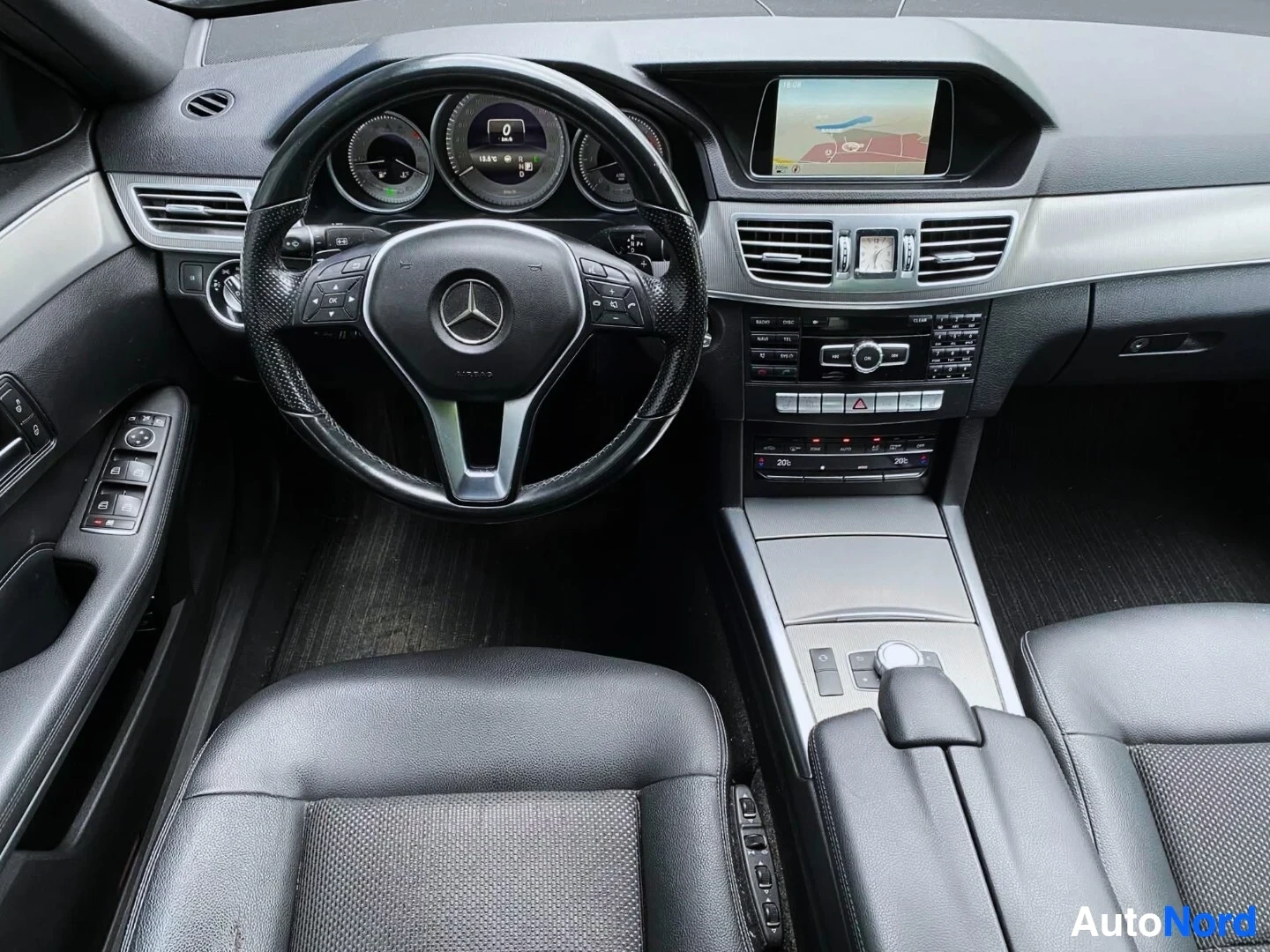 Etuistuimet: Auton ohjaustaulun etunäkymä, keskellä on Mercedes-Benz logo ohjauspyörässä, tummat ratti ja instrumentit, valkoinen hallintapaneeli, näyttöruutu keskellä näytt