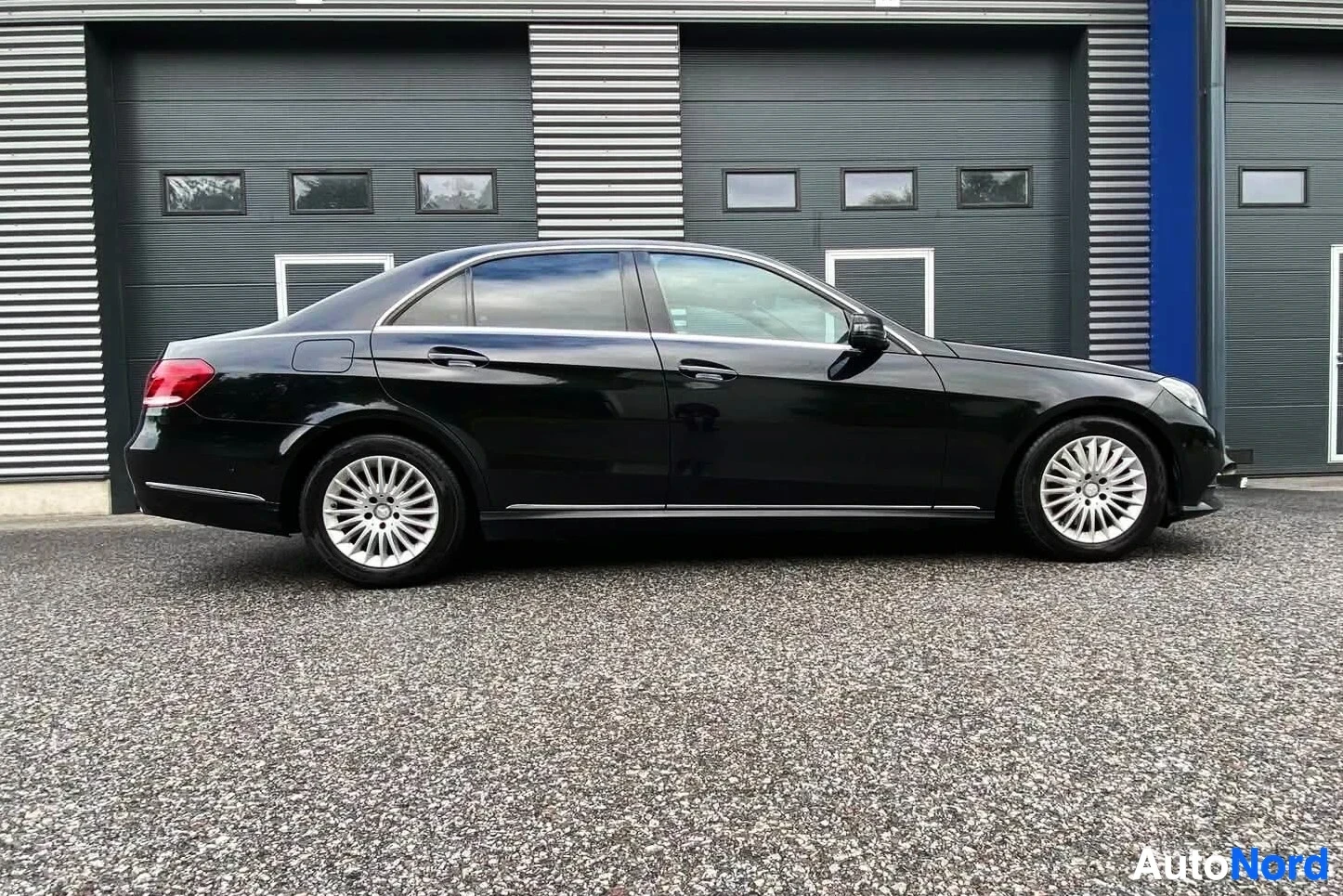 Sivulta: Musta Mercedes-Benz sedan, profiili vasemmalta oikealle, alumiini-valkoisella teollisuushallilla taustalla.