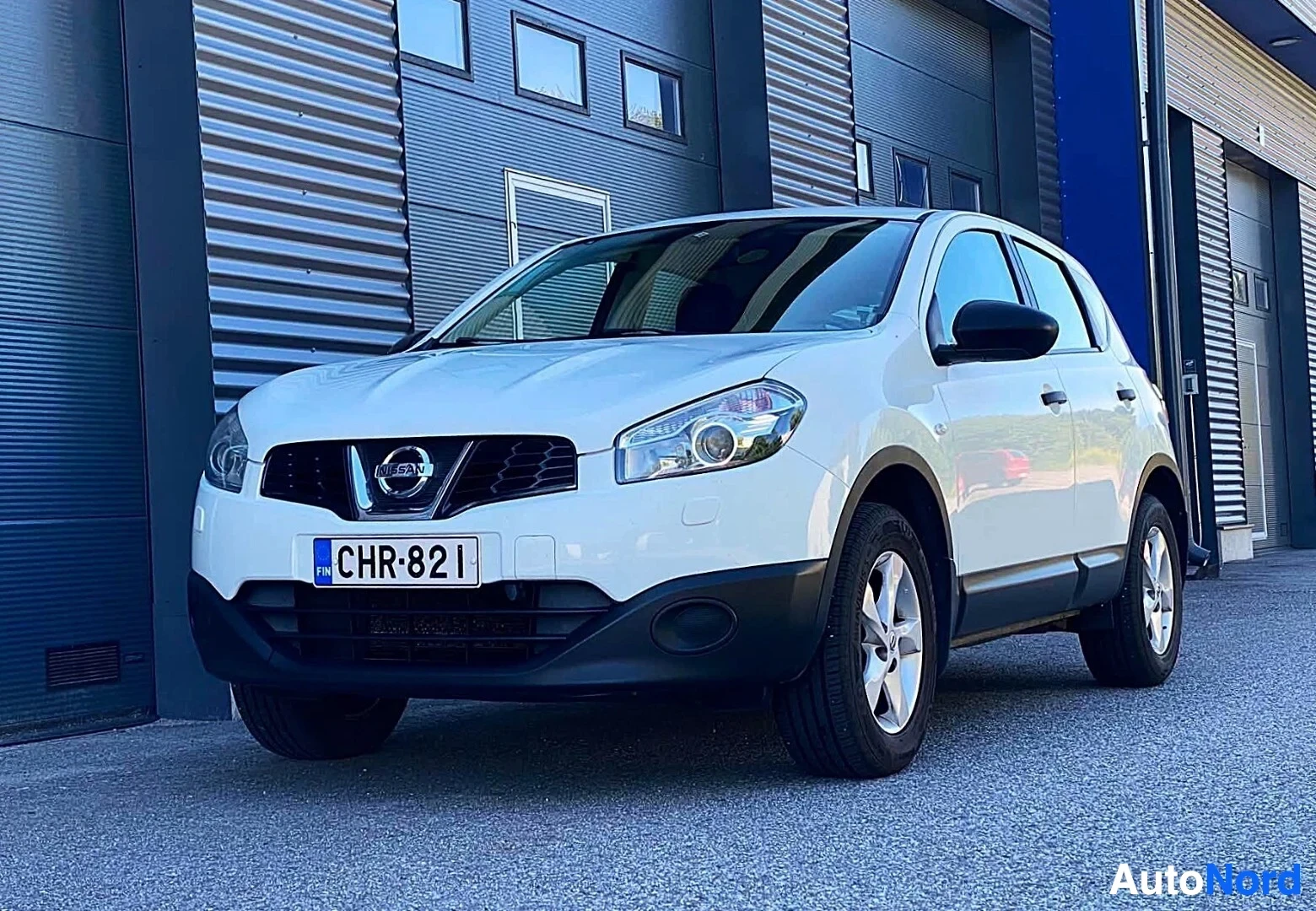 Etuviisto, nokka vasemmalle: Valkoinen Nissan Qashqai maastoauto näkyy vasemmalta etukulman näkökulmasta sinisellä teollisuushallien taustalla. Rekisterinumero on näkyvissä
