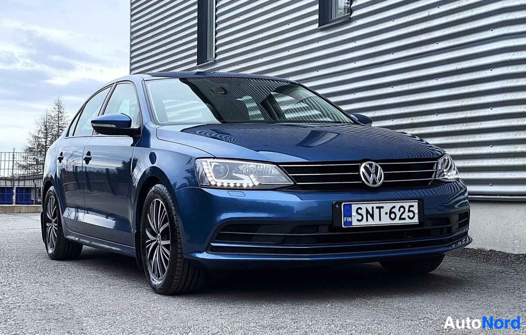 Etuviisto, nokka vasemmalle: Sininen Volkswagen Jetta sedan edestä kolmen neljäsosan kulmasta, rekisterinumero, metalliseen rakennukseen vasten