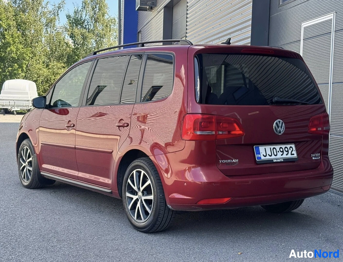 Sivulta: Punainen Volkswagen Touran sivulta kuvattuna oikealla puolella. Rekisterinumero näkyy. Vasen ovi on auki. Tekstit "TOURAN" ja "2.0 TDI" näkyvät takakont