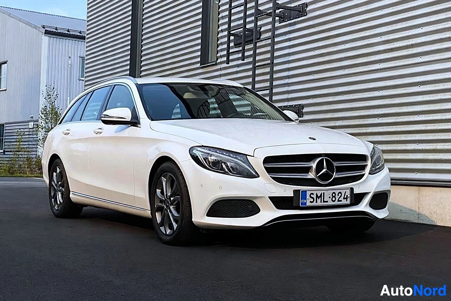 Etuviisto, nokka oikealle: Edessä oikealle päin kuvattu valkoinen Mercedes-Benz C-sarjan farmariauto, rekisterinumero, parkissa rakennuksen edessä