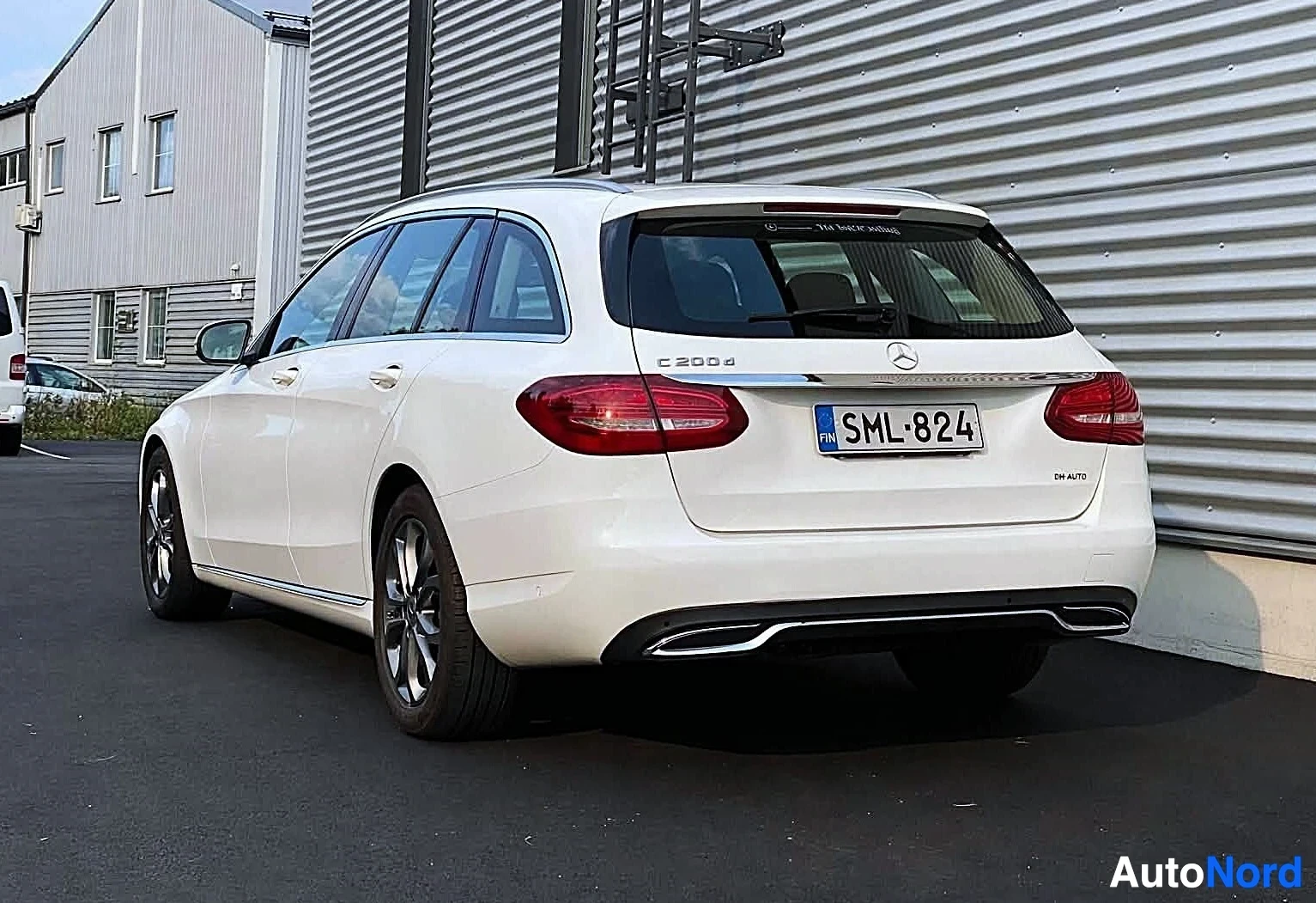 Takaviisto: Takana vasemmalle päin kuvattu valkoinen Mercedes-Benz C-sarjan farmariauto, rekisterinumero, parkissa rakennuksen edessä. Takaluukussa merkinnät 'C 200