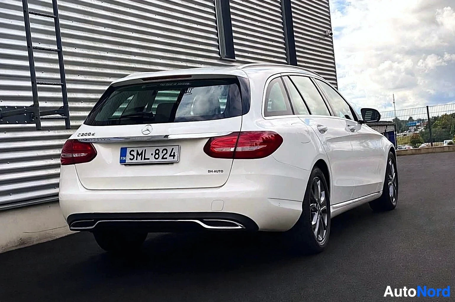 Takaviisto: Takana vasemmalle päin kuvattu valkoinen Mercedes-Benz C-sarjan farmariauto, rekisterinumero, parkissa rakennuksen edessä. Takaluukussa merkinnät 'C 200