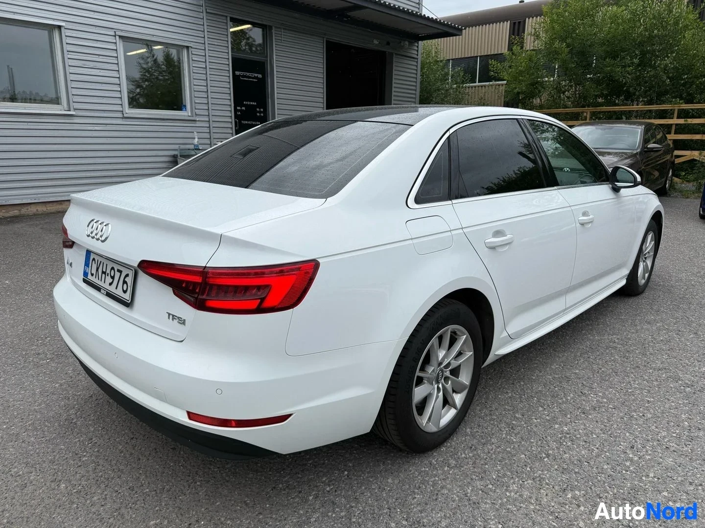 Takaviisto: Valkoinen Audi A4 sedan, kuvattu takakolmen neljänneksen kulmasta, pysäköitynä hallin edustalle.