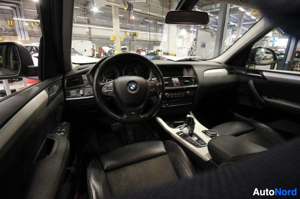 Ohjaamo: Auton sisäosa edestä, musta nahkainen penkki, mustat nahkaiset turvatyynyt sivuilla, musta ratti BMW-logolla, harmaa keskusnäyttö.