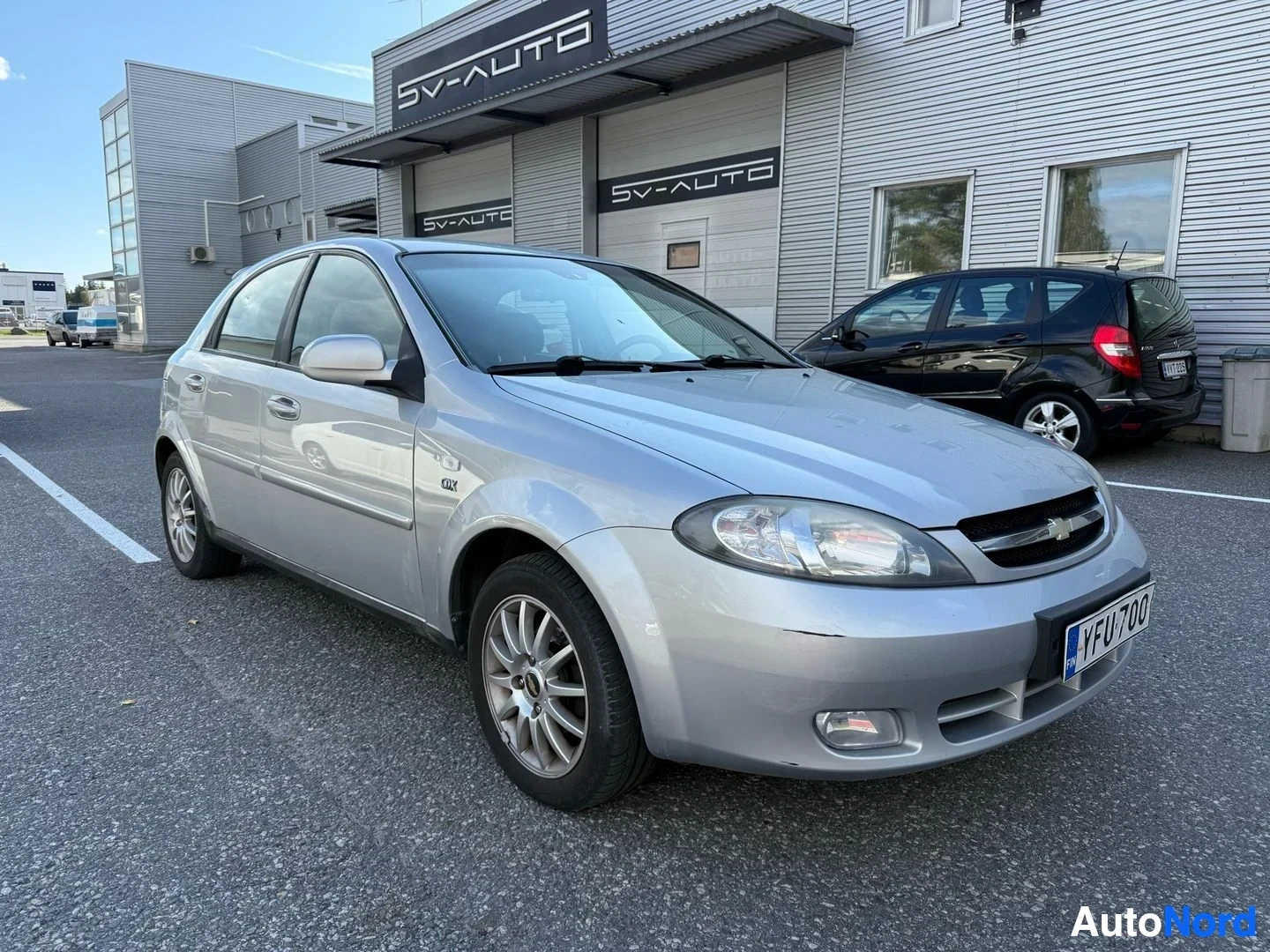 Etuviisto, nokka vasemmalle: Hopeinen Chevrolet Lacetti edestä vasemmalle, pysäköity autotallien edustalla, rekisterinumero YFU700 näkyy.