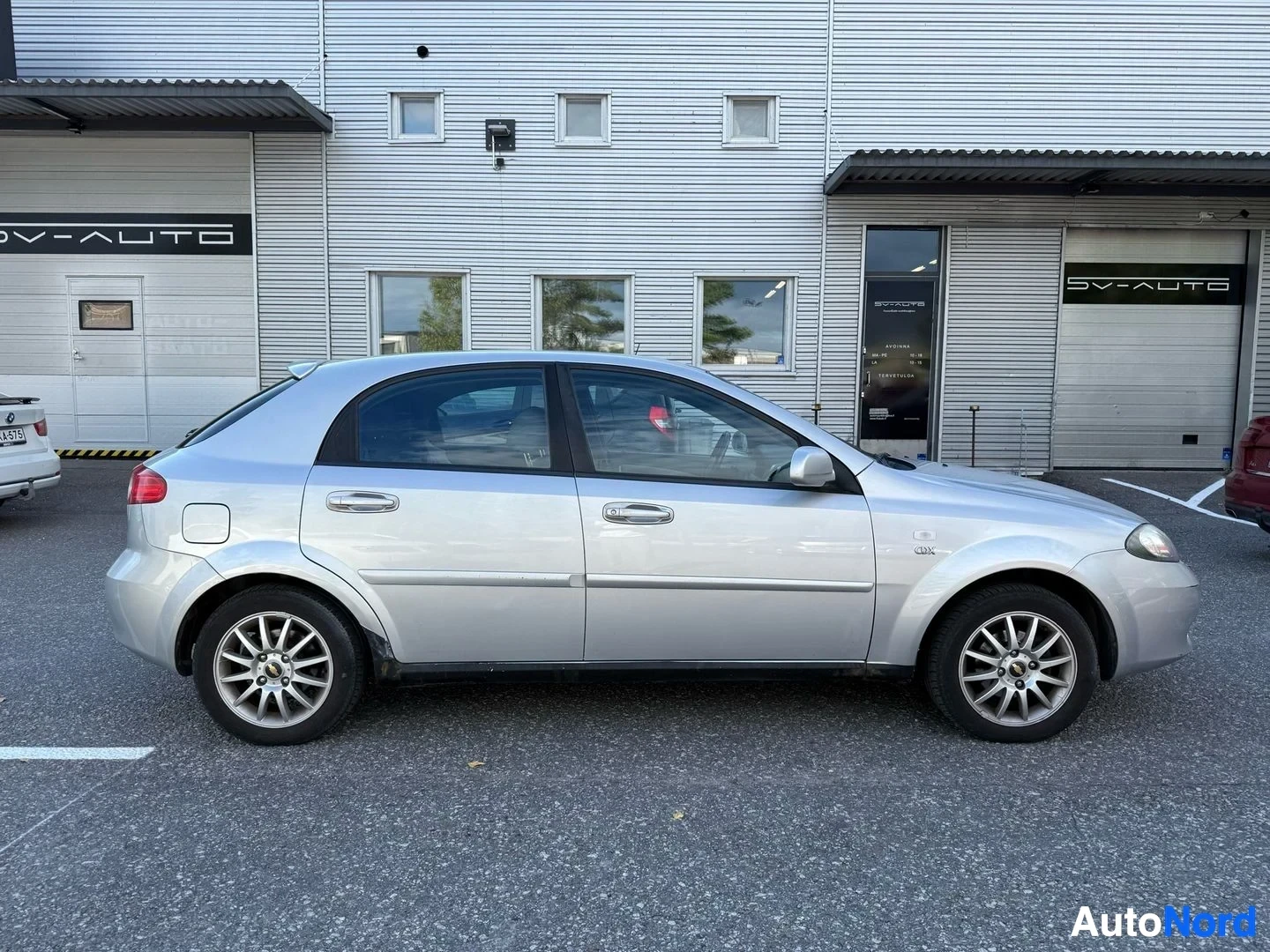 Sivulta: Hopeinen Chevrolet Lacetti sivusta, pysäköity autotallien edustalla, kaikki neljä ovea näkyvät.