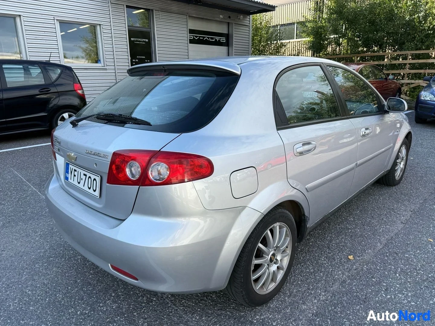 Takaa: Hopeinen Chevrolet Lacetti takaa, pysäköity autotallien edustalla, rekisterinumero YFU700 näkyy, punaiset takavalot näkyvissä.