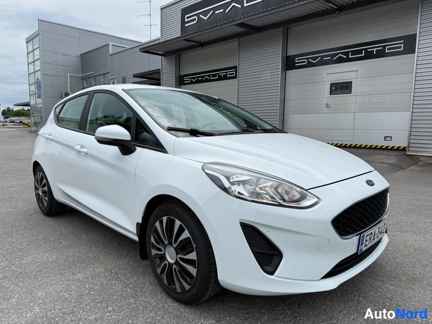 Etuviisto, nokka vasemmalle: Valkoinen Ford Fiesta etupuoli sivulta näkymä SV-AUTO liikkeen edessä, rekisterikilpi näkyvillä.