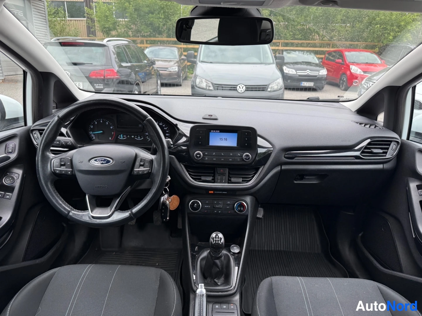 Etuistuimet: Ford Fiestan sisätila etupuolelta, rata Ford-logolla, sininen näyttö kojelaudalla aika 15:16, harmaa istuin, manuaalivaihteisto, parkkipaikka taustalla
