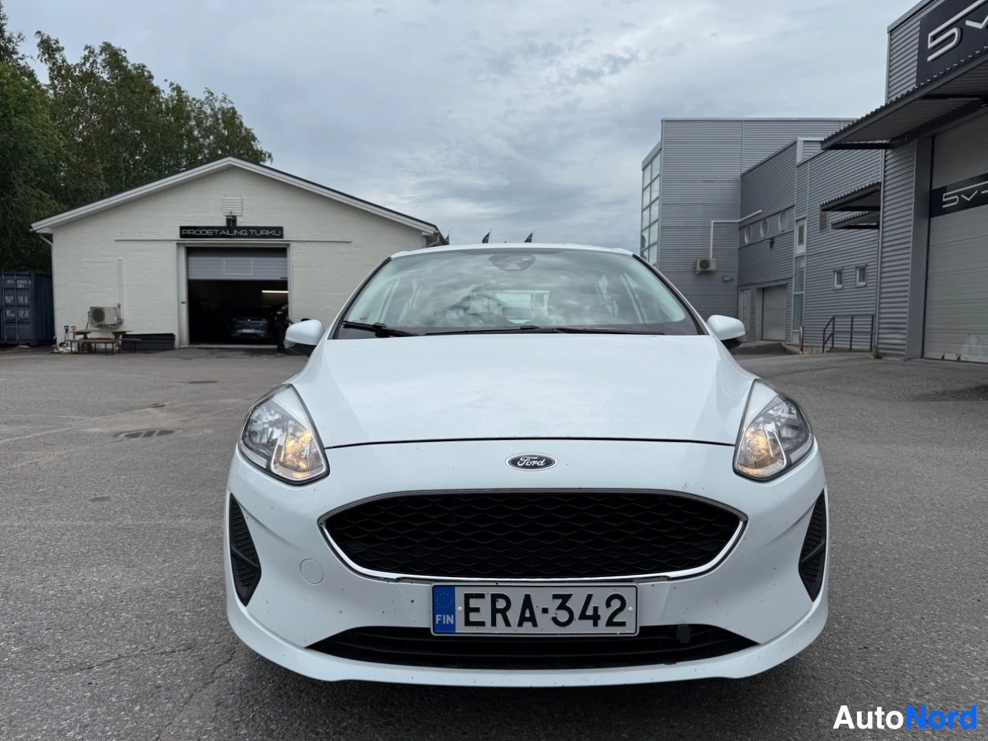 Edestä: Valkoinen Ford Fiesta etupuoli suoraan kuvattuna, rekisterinumero, Ford-merkki ja mustat ajovalot näkyvissä