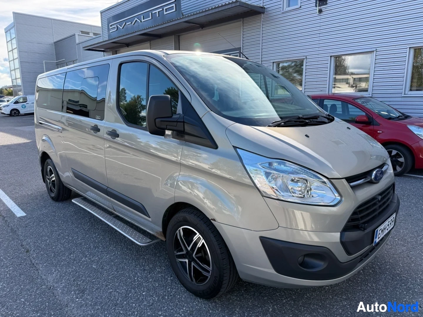Etuviisto, nokka oikealle: Valkoinen Ford Tourneo Custom -pakettiauto kuvattu kolmen neljänneksen kulmasta vasemmalta oikealle, pysäköitynä autotallilla edessä