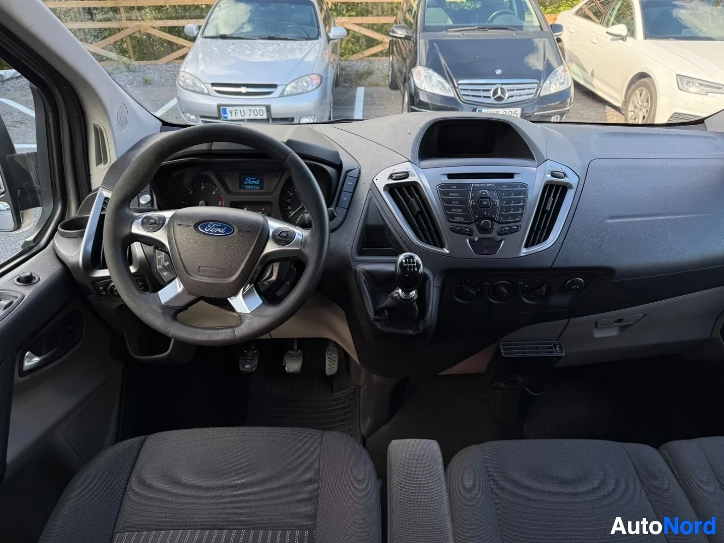 Etuistuimet: Ford Tourneo Custom sisätila samasta kulmasta, Ford-logolla varustettu mustavalkoinen kahvalla varustettu monikäyttöratti, siniset värimittarit, keskikonsoli il