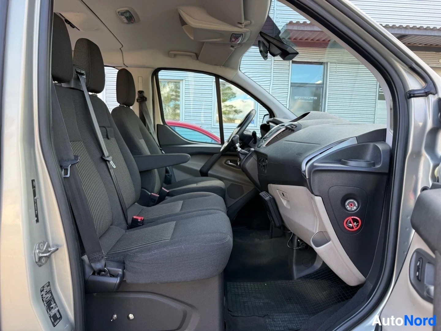 Etuistuimet: Ford Tourneo Custom sisätila sivusta, kuljettaja- ja matkustajaistuin tumman kankaalla verhoilluina, kuljettajan ratti, keskikonsoli ja kojelautapaneeli, oikeal