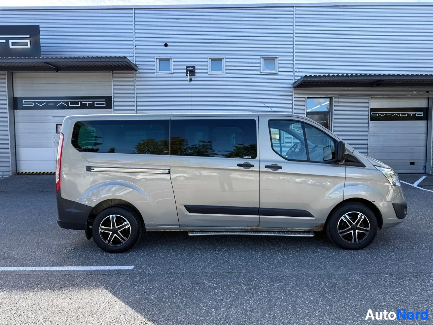 Sivulta: Valkoinen Ford Tourneo Custom -pakettiauto sivukuvassa vasemmalle päin, pysäköitynä siniseinäisen hallirakenteen edessä