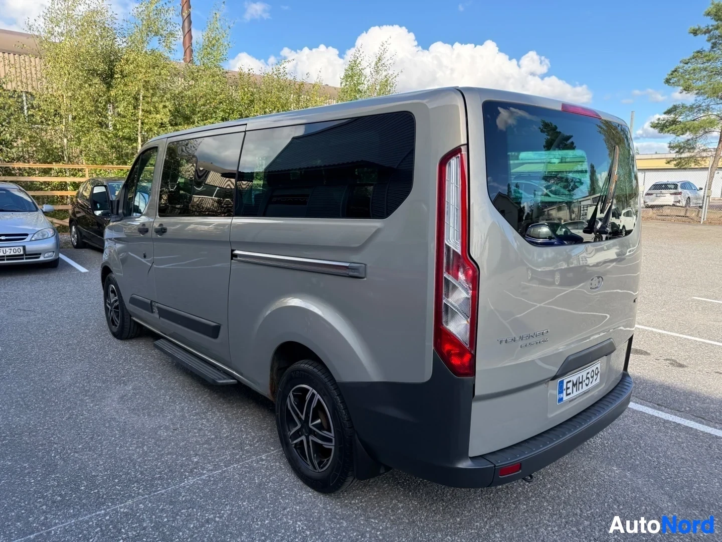 Takaviisto: Hopeanvärinen Ford Tourneo Custom -pakettiauto näkyy takavasemmalta, pysäköitynä. Näkyvissä rekisterikilpi, punaiset takavalaisimet ja hopeanvärinen run
