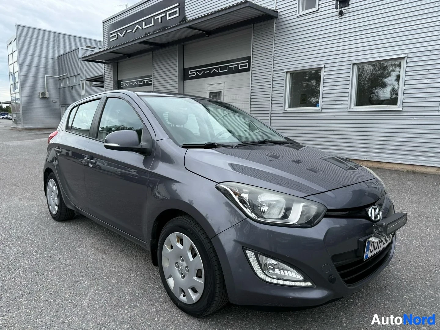 Etuviisto, nokka oikealle: Harmaa Hyundai i20 etulta kolmen neljäsosan kulmasta autotallin edessä, vasen etulovi auki