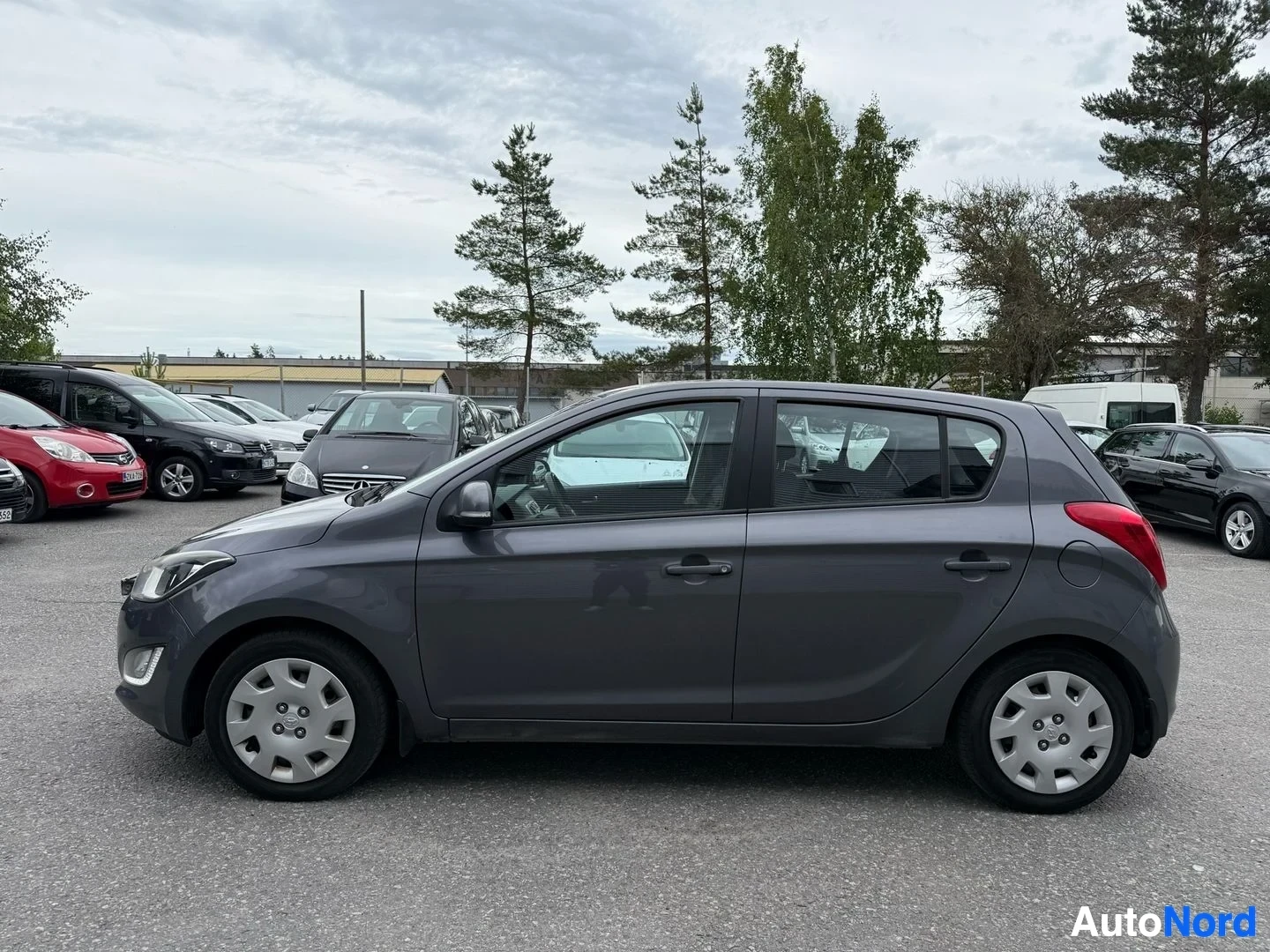 Sivulta: Harmaa Hyundai i20 oikealta sivulta nähtynä, valkoiset vanteet ja punaiset takavalot näkyvissä.