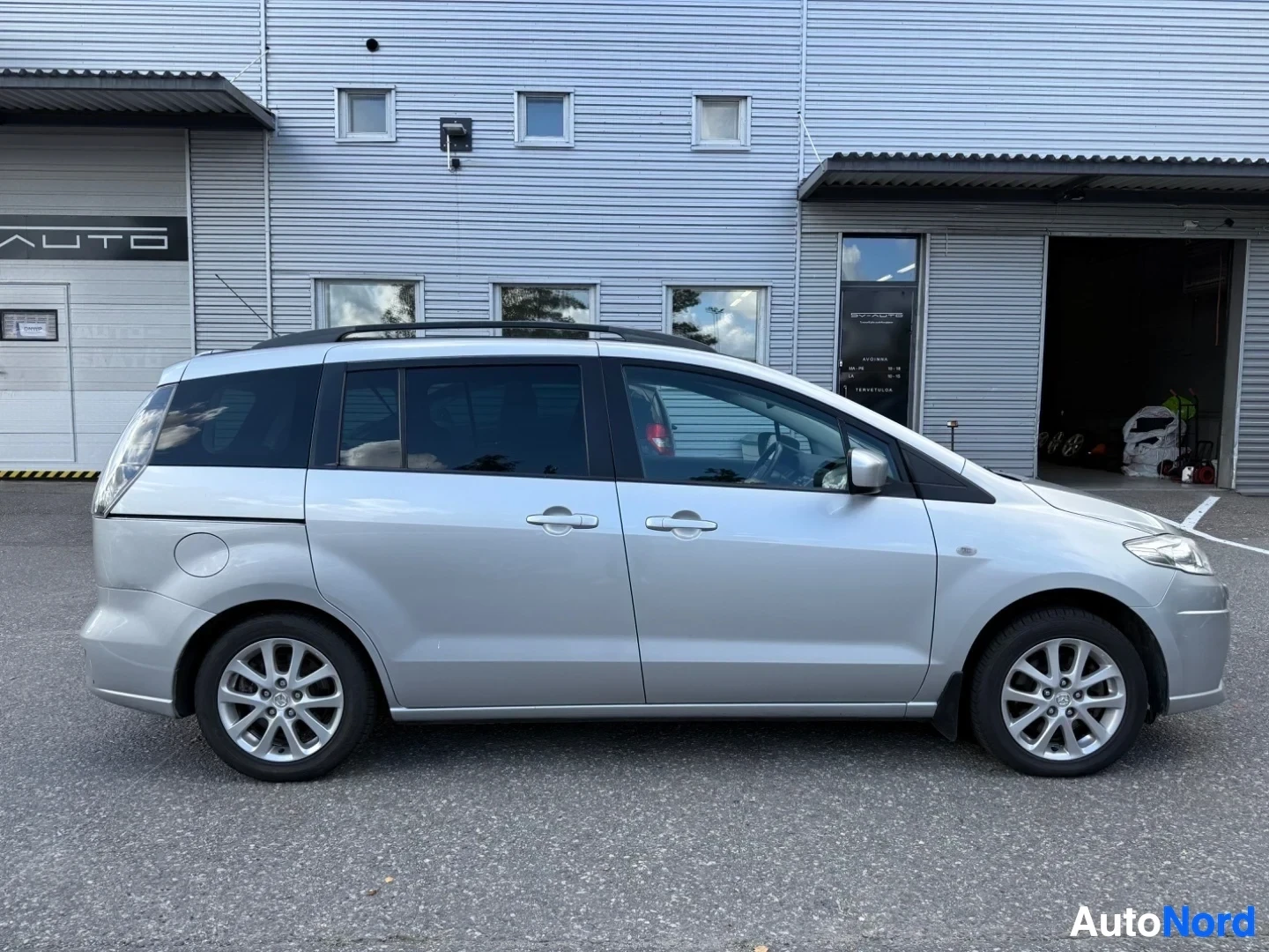 Sivulta: Hopeanvärinen Mazda-monikäyttöauto profiilista oikealle kuvattu, teollisuusrakennuksen edessä pysäköity