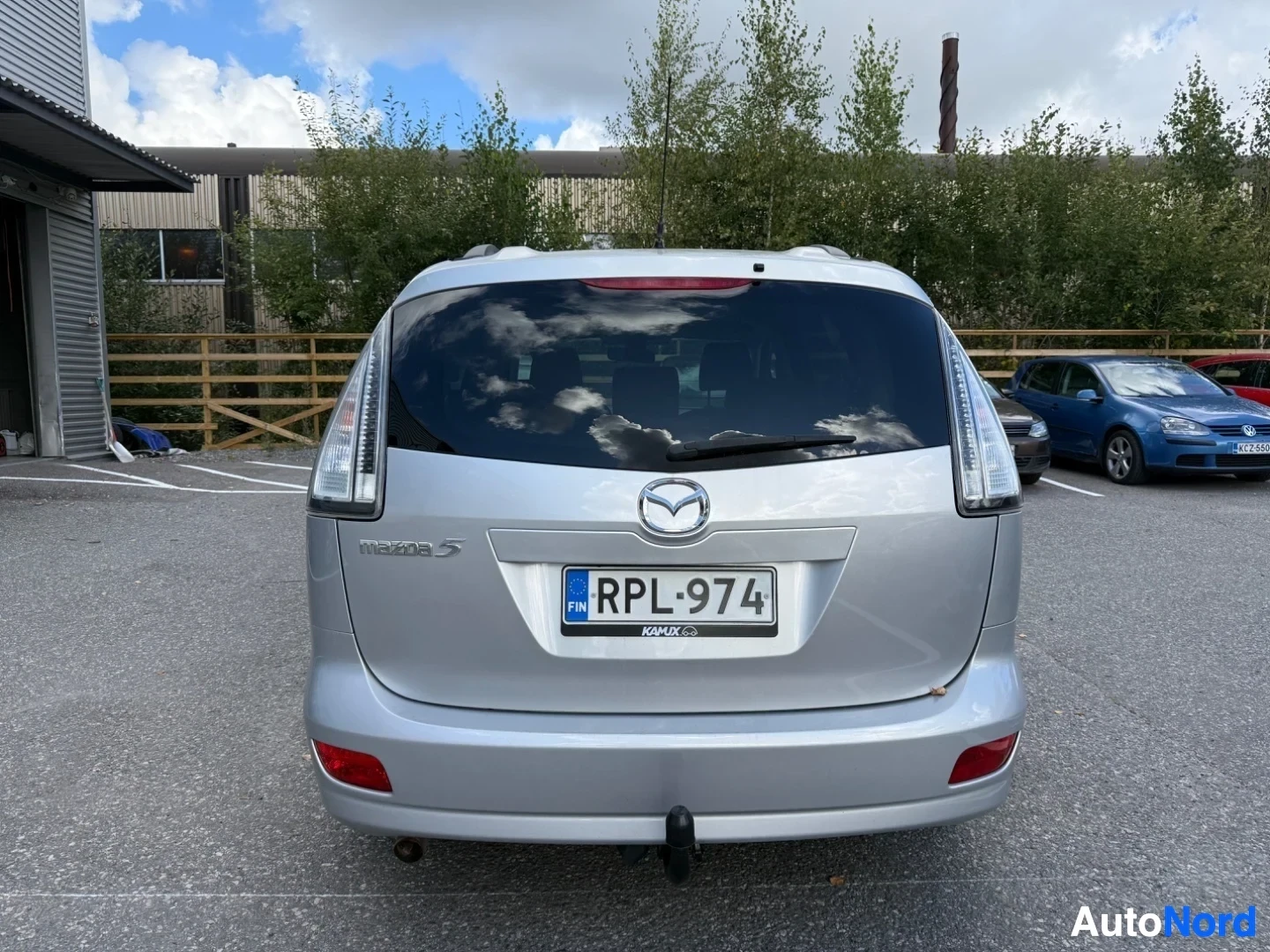 Takaa: Hopeanvärinen Mazda 5 -monikäyttöauto takaosasta kuvattu, taka-ovi, perävalot ja rekisterikilpi näkyvät, puinen aita taustalla