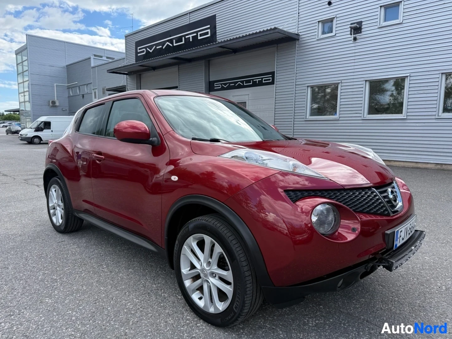 Etuviisto, nokka vasemmalle: Punainen Nissan Juke, kuvattu kolmen neljänneksen kulmasta vasemmalta. Näkyvissä koko etupuoli, oikea sivu ja osa takaosasta. Valkoisen kehyksen pyörät