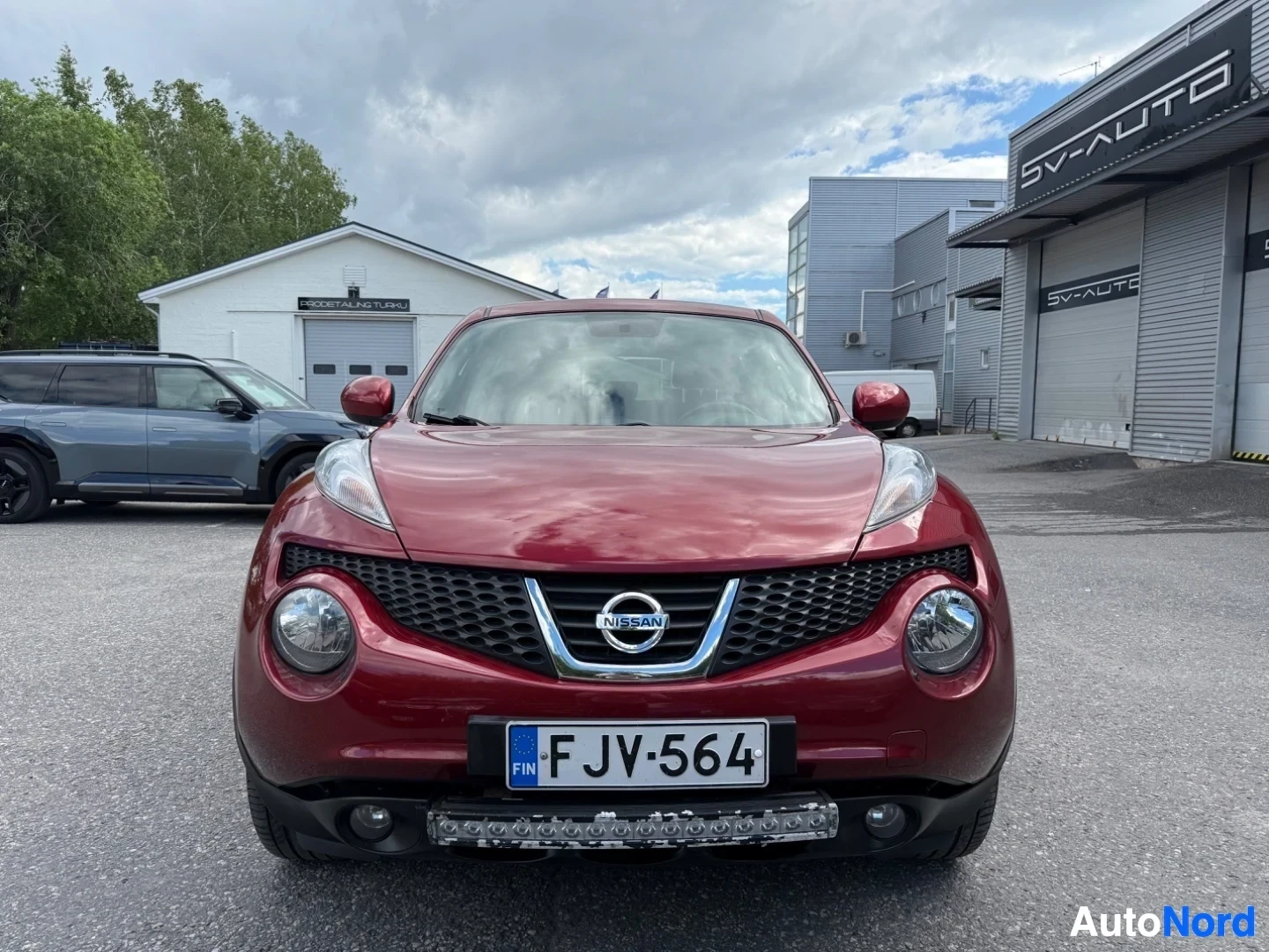Edestä: Punainen Nissan Juke edestä kuvattuna. Näkyy Nissan-merkki, mustan väriset säleikkökaari, LED-ajovalot, rekisterikilpi FJV564 ja LED-ajovalopalkki.