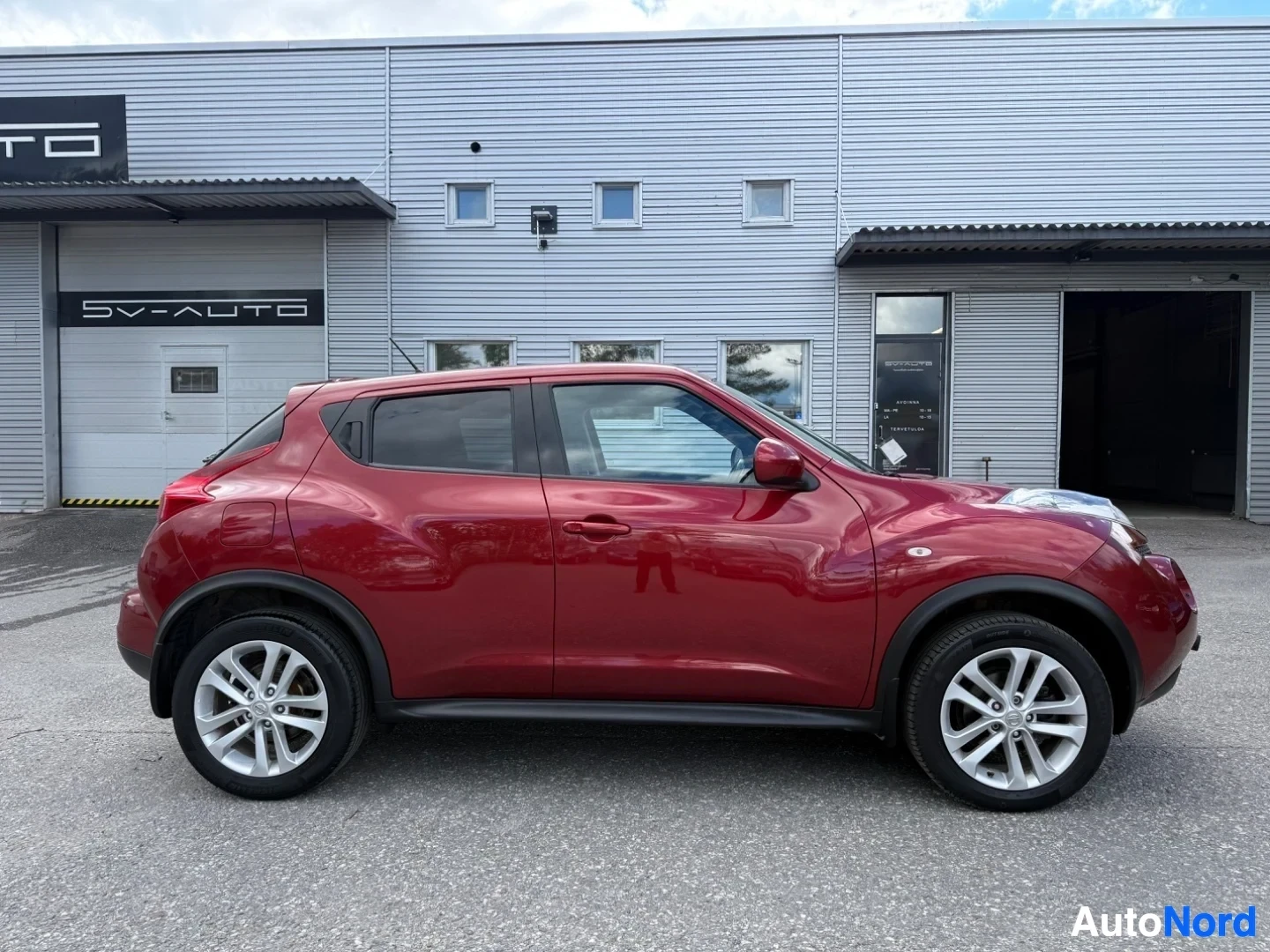 Sivulta: Punainen Nissan Juke, kuvattu oikealta sivulta. Näkyvissä koko oikea sivu, molemmat ovet ja pyörät. Valkoisen kehyksen pyörät. SV-AUTO huoltoasema näkyy taustal