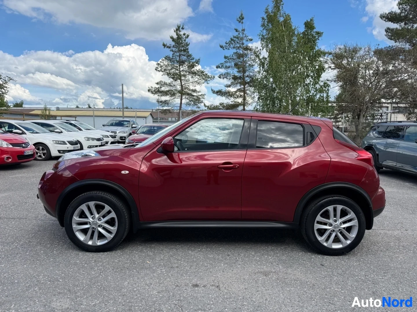 Sivulta: Punainen Nissan Juke vasemmalta sivulta kuvattuna. Näkyy valkoisen väriset vanteet ja koko auton profiili.